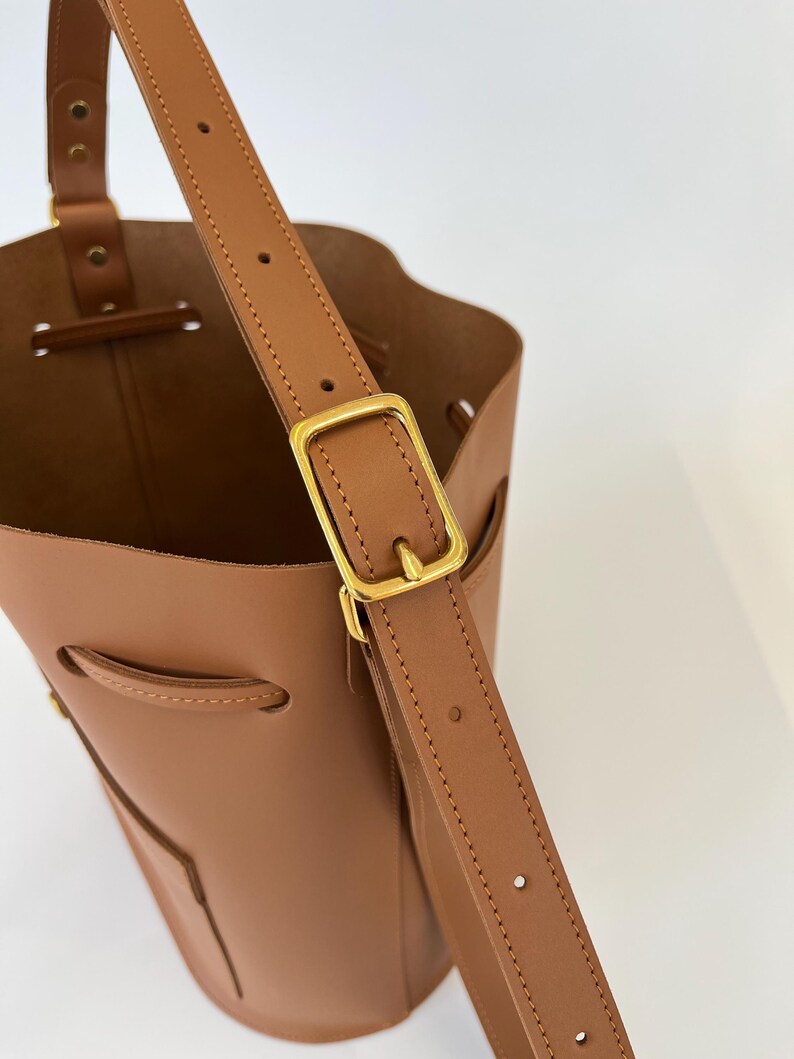 K&ouml;nnte beinhalten: Eine braune Ledereimer-Tasche mit einer goldenen Schnalle und einem verstellbaren Trageriemen. Die Tasche hat einen Kordelverschluss und eine kleine Tasche auf der Vorderseite.