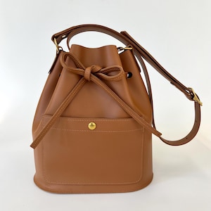 Tan leather bucket bag with a front pocket secured by a gold snap button, a drawstring closure tied in a bow, and an adjustable shoulder strap with gold hardware