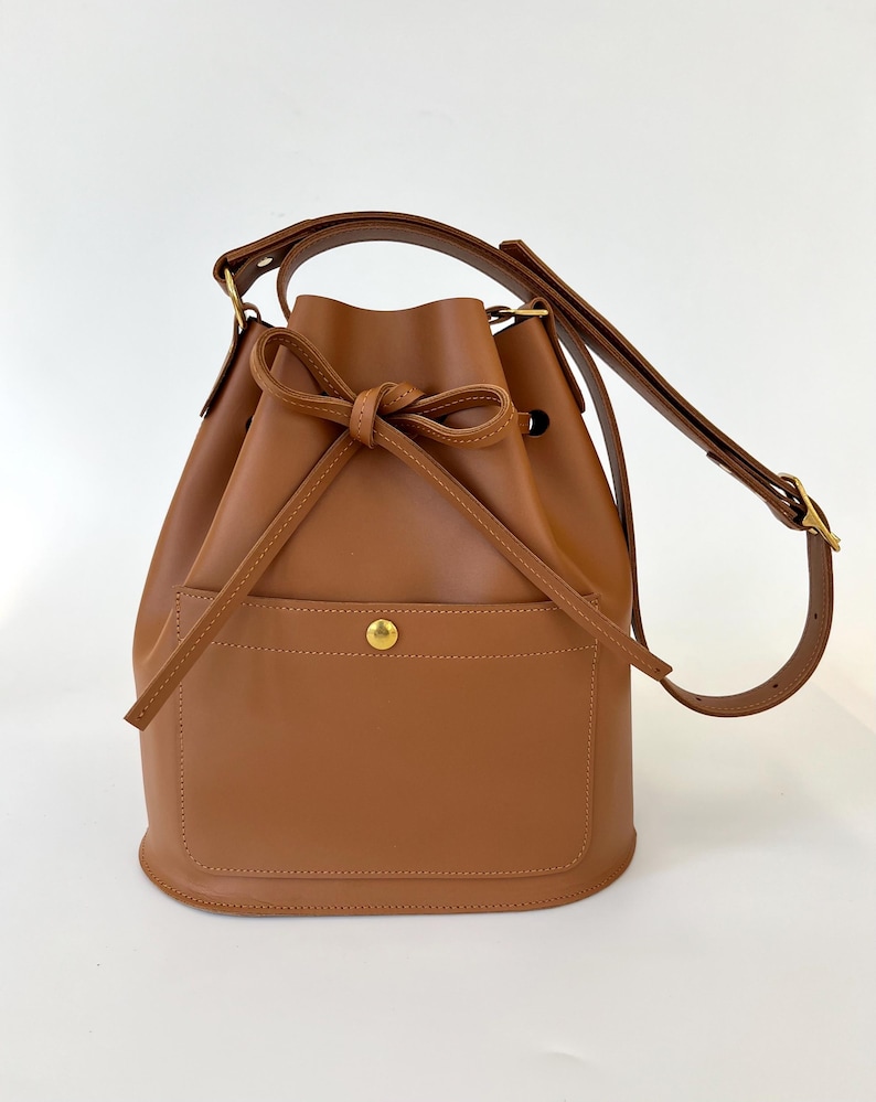 Tan leather bucket bag with a front pocket secured by a gold snap button, a drawstring closure tied in a bow, and an adjustable shoulder strap with gold hardware