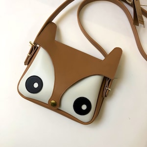 Fox face leather bag. Square shaped cartoon minimalistic fox head. white face with black eyes and brown ears and nose panel that close the bag at the front. leather adjustable strap. crossbody bag. One of La Lisette's best sellers!