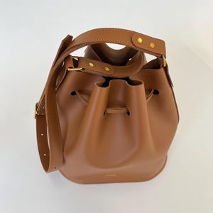 K&ouml;nnte beinhalten: Eine braune Ledereimer-Tasche mit goldenen Beschl&auml;gen und einem Kordelzug.