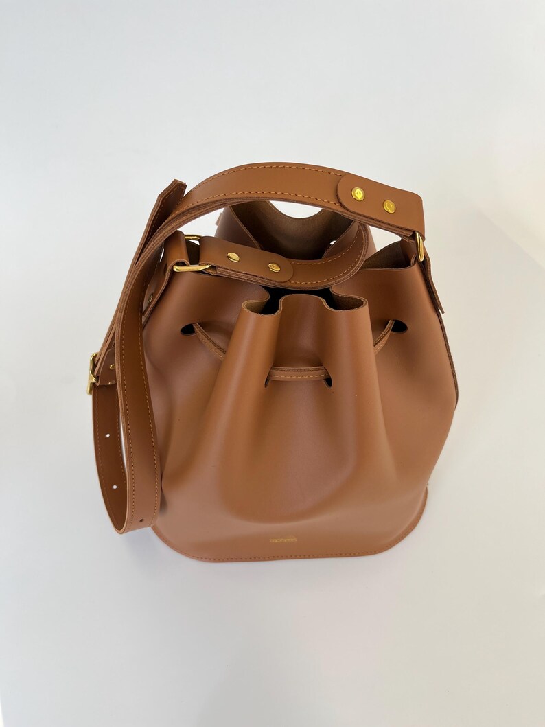 K&ouml;nnte beinhalten: Eine braune Ledereimer-Tasche mit goldenen Beschl&auml;gen und einem Kordelzug.