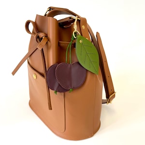 K&ouml;nnte beinhalten: Eine braune Ledereimer-Tasche mit Kordelzug und abnehmbarem Trageriemen. Die Tasche hat eine kleine Tasche auf der Vorderseite und ist mit einem gr&uuml;nen Lederblatt und zwei burgunderfarbenen Lederkirschen verziert.