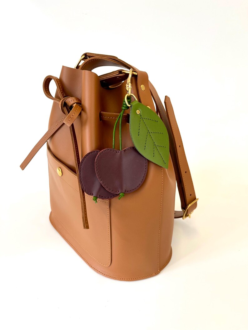 K&ouml;nnte beinhalten: Eine braune Ledereimer-Tasche mit Kordelzug und abnehmbarem Trageriemen. Die Tasche hat eine kleine Tasche auf der Vorderseite und ist mit einem gr&uuml;nen Lederblatt und zwei burgunderfarbenen Lederkirschen verziert.