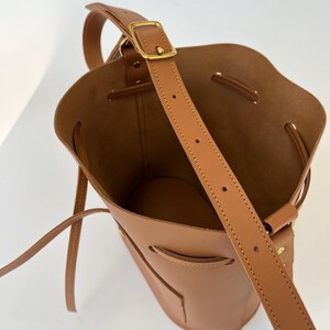 K&ouml;nnte beinhalten: Eine braune Ledereimer-Tasche mit einem langen Riemen und einer goldenen Schnalle. Die Tasche hat einen Kordelverschluss und eine kleine Tasche auf der Vorderseite.