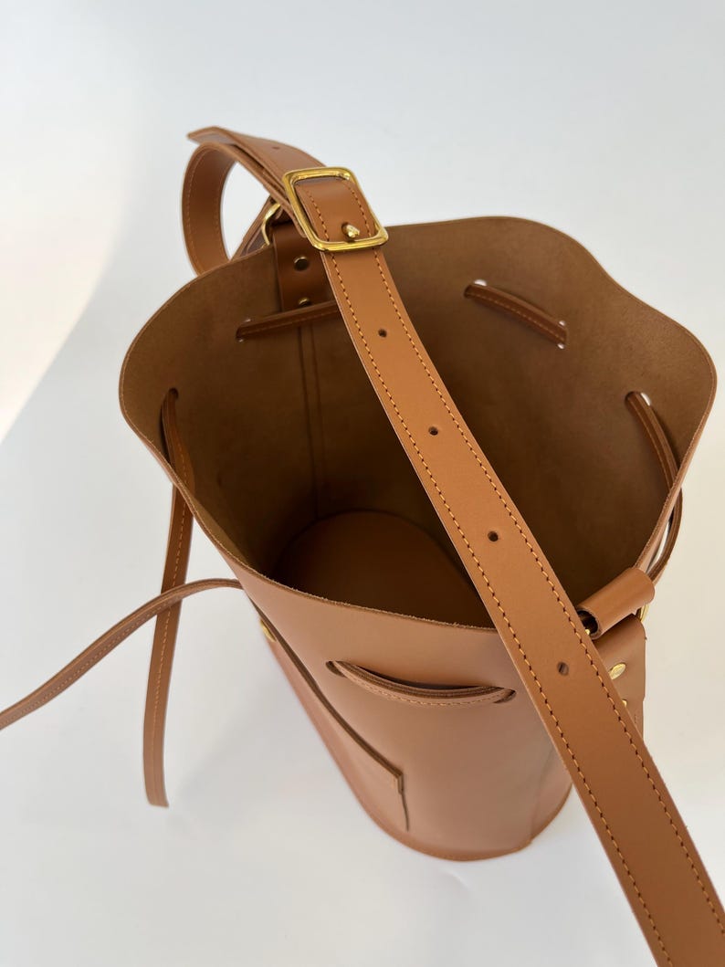 K&ouml;nnte beinhalten: Eine braune Ledereimer-Tasche mit einem langen Riemen und einer goldenen Schnalle. Die Tasche hat einen Kordelverschluss und eine kleine Tasche auf der Vorderseite.