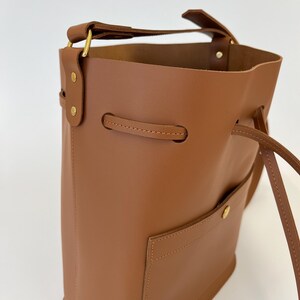 K&ouml;nnte beinhalten: Eine braune Ledereimer-Tasche mit einem Henkel oben und einem Kordelzugverschluss. Die Tasche hat eine kleine Tasche auf der Vorderseite und eine goldfarbene Schnalle am Henkel.