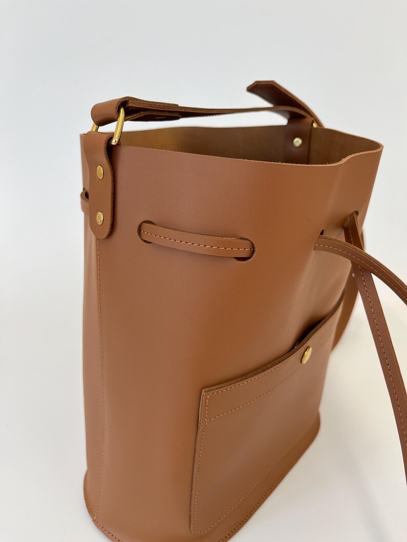 K&ouml;nnte beinhalten: Eine braune Ledereimer-Tasche mit einem Henkel oben und einem Kordelzugverschluss. Die Tasche hat eine kleine Tasche auf der Vorderseite und eine goldfarbene Schnalle am Henkel.