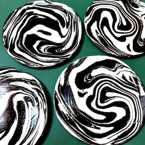 May include: Four round coasters with a black and white swirled pattern. The coasters have a glossy finish and a unique, marbled design. The coasters are arranged on a green surface.