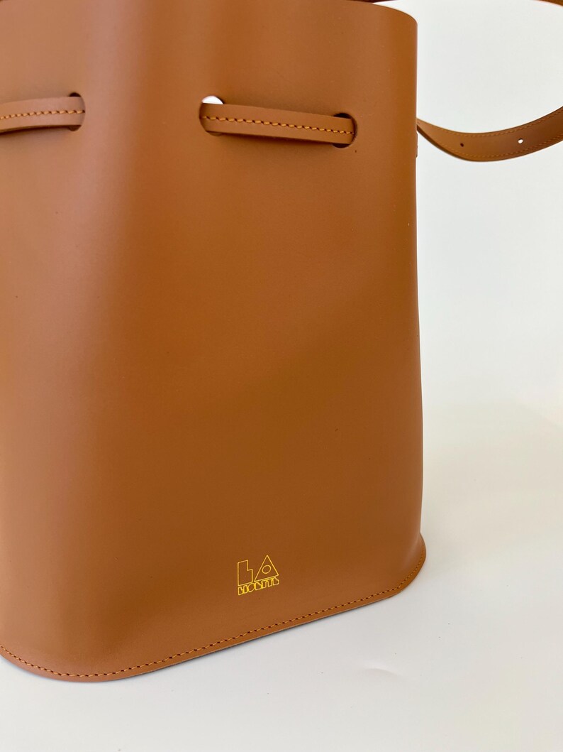 K&ouml;nnte beinhalten: Eine braune Ledereimer-Tasche mit einem goldenen Logo, auf dem "LA BAGUETTE" steht.