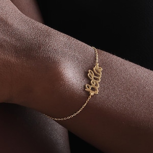 May include: A delicate gold bracelet spelling out the name "Bella" in cursive script. The bracelet is attached to a thin chain and is worn on a person's wrist. The background is a dark, solid colour.