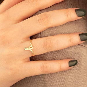 Puede incluir: Un delicado anillo de oro con un diseño de letra "J" en cursiva. El anillo es delgado y se coloca en un dedo, mostrando su estilo minimalista. El color dorado del anillo contrasta con el tono de la piel y la ropa de tonos neutros en el fondo.