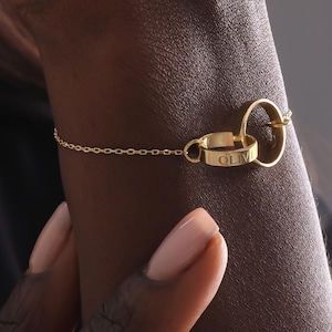 May include: A gold-colored bracelet with two interlocking rings and a delicate chain. One ring is engraved with the name "OLIV". The bracelet is worn on a wrist with manicured nails, against a dark background.