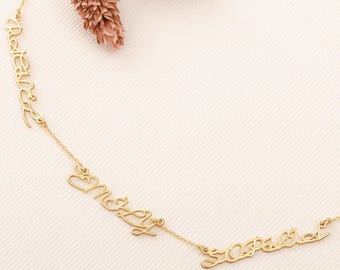 May include: A delicate gold-colored necklace featuring three names in a cursive font. The names are connected by a fine chain. The necklace is displayed on a light-colored surface, with a decorative element in the background.
