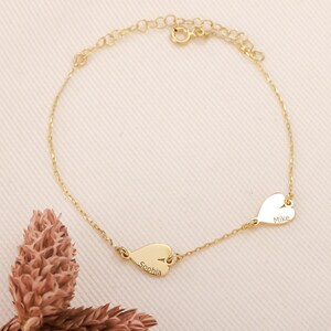 May include: A delicate gold-colored bracelet featuring two heart-shaped charms. One charm is engraved with the name "Sophia," and the other with "Mike." The bracelet is on a light pink surface.