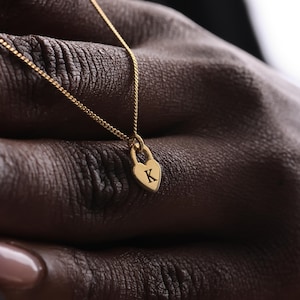 Op de afbeelding: Een goudkleurige ketting met een hartvormige hanger met de gegraveerde letter "K". De hanger is ontworpen om op een hangslot te lijken. De ketting is een delicate gourmette-stijl.