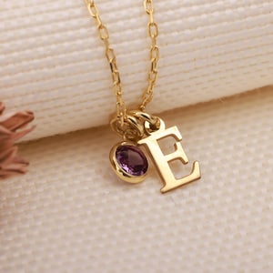 Peut inclure: Un collier doré avec un pendentif en forme de lettre "E" et une petite pierre précieuse ovale violette. La chaîne et le pendentif sont présentés sur un fond blanc cassé.