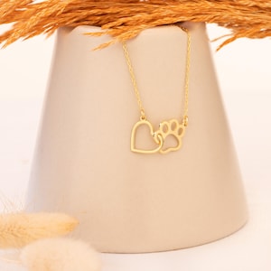 May include: A gold-colored necklace featuring a heart and a paw print pendant. The pendant is connected to a delicate chain. The necklace is displayed against a neutral background, with dried ornamental grass.