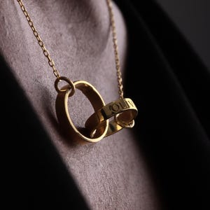 May include: A gold-colored necklace featuring two interlocking rings. One ring is engraved with the name "OLIV" in capital letters. The necklace hangs against a dark background, highlighting the jewelry's design.