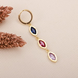 May include: A gold-colored earring with a hoop and a dangling design. The dangling portion features three marquise-shaped gemstones in blue, red, and purple. The earring is set against a neutral background.