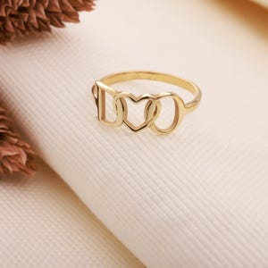 Może przedstawiać: Złoty pierścionek z wyciętymi literami "I", sercem i "O". Pierścionek jest umieszczony na białym tle. Pierścionek ma prosty, elegancki design.