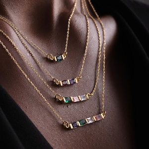 May include: Four gold necklaces with colorful gemstone bars. Each necklace features a delicate gold chain and a bar pendant with emerald green, ruby red, amethyst purple, and pink gemstones. The necklaces are layered for a stylish look.