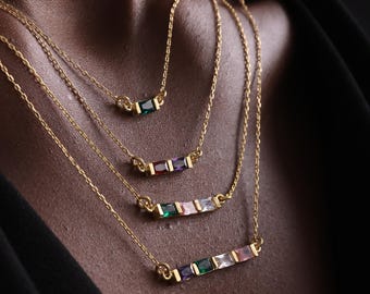 Op de afbeelding: Vier gouden kettingen met gekleurde edelsteen staven. Elke ketting heeft een fijne gouden ketting en een staafhanger met smaragdgroene, robijnrode, amethistpaarse en roze edelstenen. De kettingen zijn gelaagd.