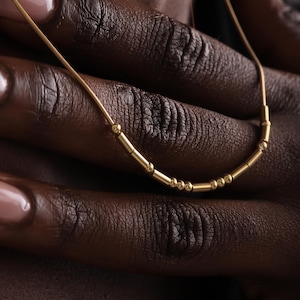 以下が含まれることがあります： 小さな円筒形と球形のビーズが連なった繊細なゴールドネックレス。暗い背景を背景に、明るいメタリック仕上げを強調しています。このジュエリーは、シンプルでエレガントなアクセサリーです。