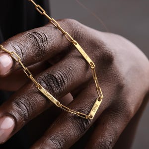 May include: A gold-colored chain necklace with rectangular nameplates. The nameplates are engraved with the names "Thomas", "Sophia", and "Olivia". The necklace is displayed against a dark background.