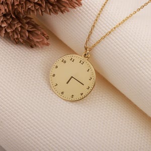 May include: A gold-coloured necklace with a circular pendant designed to look like a clock face. The pendant has black clock hands and numbers around the edge. The necklace is displayed on a white surface.
