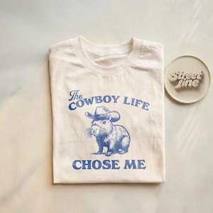May include: Cream-colored t-shirt with a blue graphic of a capybara wearing a cowboy hat. The text "The Cowboy Life Chose Me" is printed above and below the illustration. A small, round, off-white coaster with the text "Street Line" is to the right.