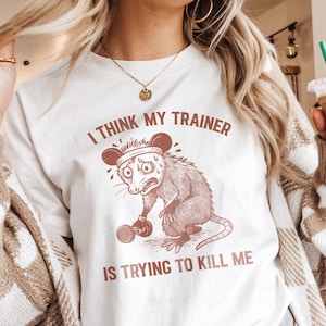 Puede incluir: Camiseta color crema con el texto "I THINK MY TRAINER IS TRYING TO KILL ME" en marrón. La camiseta presenta una zarigüeya de dibujos animados sosteniendo una pesa, que parece estresada. La camiseta es parte de una colección de ropa.