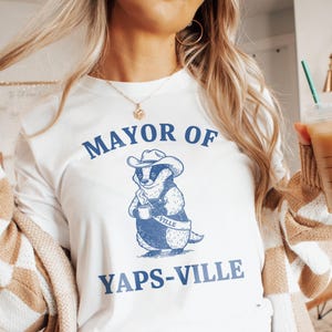 Peut inclure: T-shirt blanc à manches longues avec l'inscription bleue "MAYOR OF YAPS-VILLE". Le motif représente un blaireau de dessin animé portant un chapeau de cow-boy et une salopette, tenant une tasse de café. Le blaireau porte une écharpe avec "-VILLE".