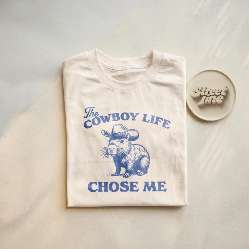 May include: Cream-colored t-shirt with a blue graphic of a capybara wearing a cowboy hat. The text "The Cowboy Life Chose Me" is printed above and below the illustration. A small, round, off-white coaster with the text "Street Line" is to the right.