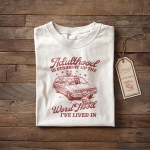 Puede incluir: Camiseta de color crema con un gráfico rojo de un coche familiar lleno de mapaches y el texto "Adulthood is straight up the Worst Hood I've lived in." Una etiqueta marrón está adjunta a la camiseta.