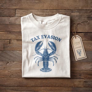 Peut inclure: T-shirt blanc cassé avec un motif de homard bleu et l'inscription "TAX EVASION" au-dessus. Une étiquette assortie avec le même motif est attachée. Le t-shirt est plié sur une surface en bois.