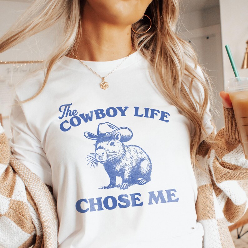 May include: White long-sleeve t-shirt with a blue graphic of a capybara wearing a cowboy hat. The text "The Cowboy Life CHOSE ME" is printed above and below the illustration. The shirt is a casual wear item.