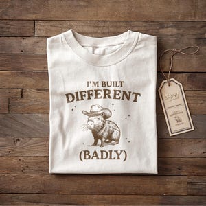 Könnte beinhalten: Cremefarbenes T-Shirt mit brauner Grafik eines Capybaras mit Cowboyhut. Der Text "I'M BUILT DIFFERENT (BADLY)" ist oberhalb und unterhalb der Illustration gedruckt. Ein braunes Etikett mit Text hängt am Shirt.