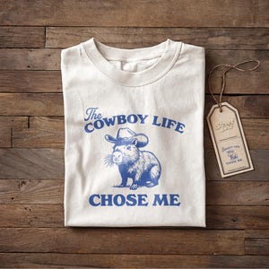 May include: Cream-colored t-shirt with the text "The Cowboy Life Chose Me" in blue, along with a blue illustration of a capybara wearing a cowboy hat. A brown tag with the text "Street Life" hangs on the side.