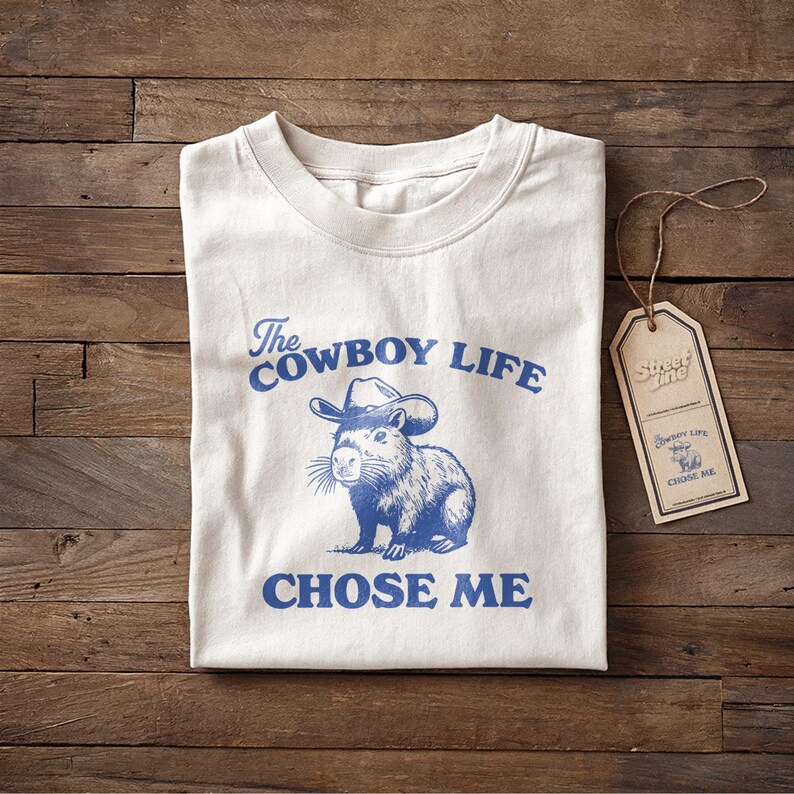 May include: Cream-colored t-shirt with the text "The Cowboy Life Chose Me" in blue, along with a blue illustration of a capybara wearing a cowboy hat. A brown tag with the text "Street Life" hangs on the side.