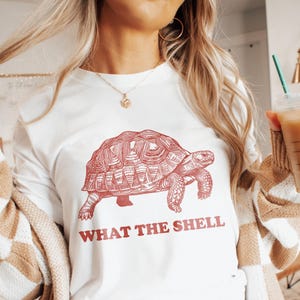 Op de afbeelding: Wit shirt met lange mouwen met een rode illustratie van een schildpad en de tekst "WHAT THE SHELL" in rode letters. Het shirt is gemaakt van een zacht materiaal.