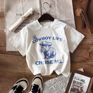 May include: White t-shirt with a blue graphic of a capybara wearing a cowboy hat and the text "The Cowboy Life Chose Me." The shirt is hanging on a hanger. Sneakers and a framed picture are in the foreground.