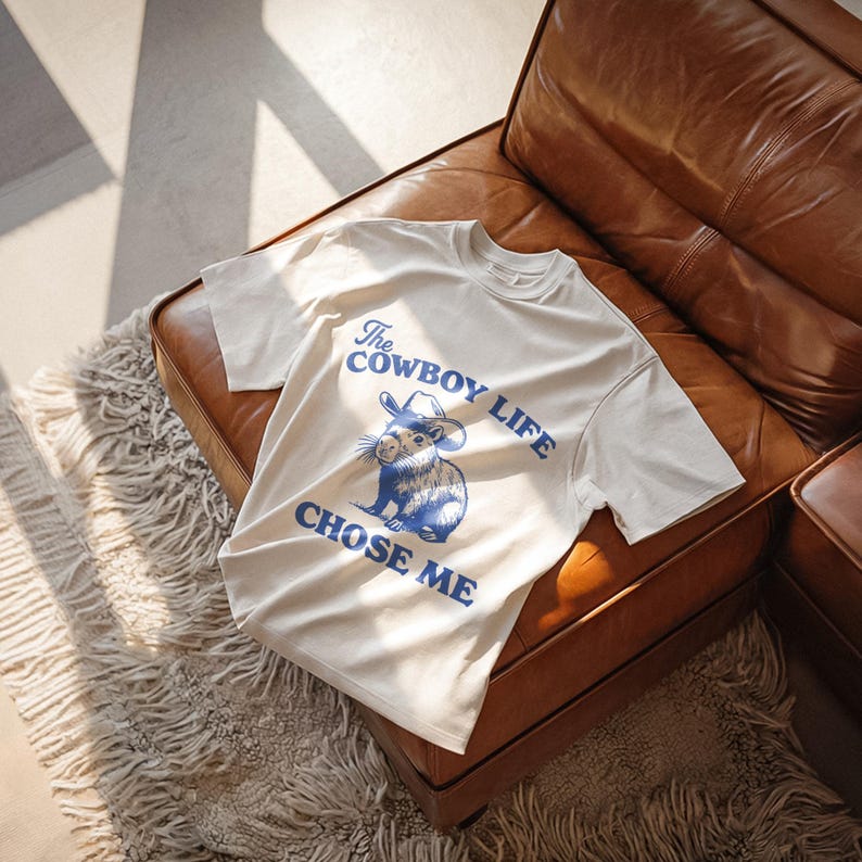 May include: Cream-colored t-shirt with the text "The Cowboy Life Chose Me" in blue, along with a blue illustration of a rodent wearing a cowboy hat. The shirt is displayed on a brown leather couch.