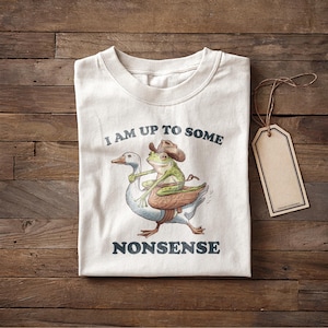 May include: Cream-colored t-shirt featuring a whimsical illustration of a goose carrying a frog wearing a cowboy hat. The text "I AM UP TO SOME NONSENSE" is printed above and below the image in dark blue.