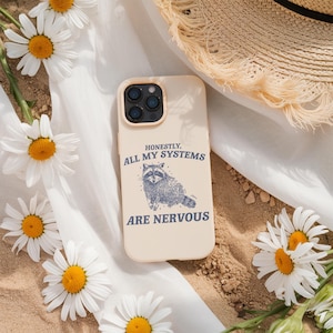 May include: A beige phone case with a raccoon illustration and the text "HONESTLY, ALL MY SYSTEMS ARE NERVOUS." The case is surrounded by daisies and a straw hat.