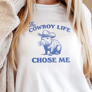 May include: White t-shirt with a blue graphic of a capybara wearing a cowboy hat. The text "The Cowboy Life CHOSE ME" is printed above and below the illustration. The shirt is made of a soft material.
