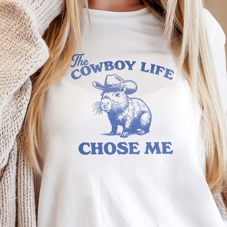 May include: White t-shirt with a blue graphic of a capybara wearing a cowboy hat. The text "The Cowboy Life CHOSE ME" is printed above and below the illustration. The shirt is made of a soft material.