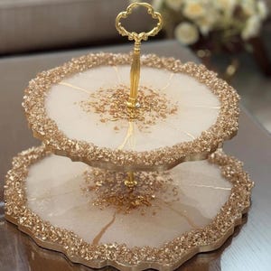 Könnte beinhalten: Ein zweistöckiger Kuchenständer mit goldenem Griff. Der Ständer ist aus weißem Harz mit goldenem Glitzer und einem Blattgold-Design.