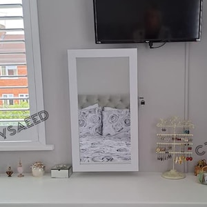 May include: A white vanity with a mirror, drawers, and a cushioned stool. The vanity has a built-in desk area and is adorned with decorative items, including perfume bottles and jewelry organizers. A flat-screen TV is mounted on the wall above.