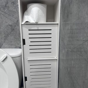 May include: A white, freestanding bathroom storage cabinet with a slatted door design. The cabinet has a shelf holding a roll of toilet paper and two bottles. The cabinet is next to a white toilet.